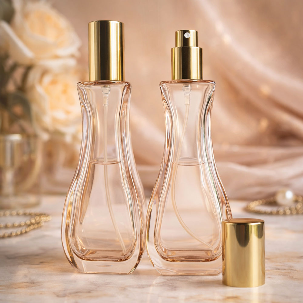 Two clear glass perfume bottles with gold spray tops displayed on a marble surface surrounded by soft pink satin, roses, and gold jewelry in warm, elegant lighting.